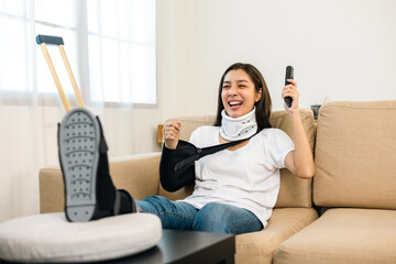 Happy woman recovery watching tv using remote from accident fracture broken bone injury with leg splints in cast neck splints collar sling support arm. Social security and health insurance concept.