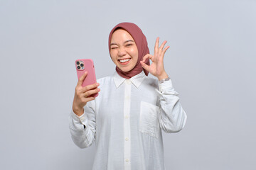 Wall Mural - Smiling young Asian Muslim woman holding mobile phone and showing okay sign isolated over white background