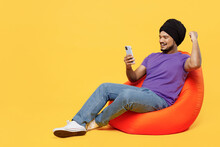 Full Body Devotee Sikh Indian Man Ties His Traditional Turban Dastar Wear Purple T-shirt Sit In Bag Chair Use Mobile Cell Phone Do Winner Gesture Isolated On Plain Yellow Background Studio Portrait.