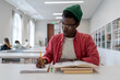 © DimaBerlin - Talented young black guy scientist wearing glasses studying in library, reading book and taking notes. Focused African American male student writing research summary, sitting at desk. Education