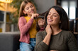 © Dorde - Photo of happy smiling school girl wearing yellow blouse and pink cardigan who is tenderly brushing mother's hair. Focus on mom who is enjoying in present moment with her daughter with eyes closed.
