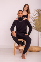 A happy beautiful , multiracial couple, in black clothes, are sitting near the wall in the room.