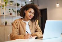 Confident African American Young Woman With Stylish Afro Hairstyle, Smart Student Studying, Taking Notes On Notepad Watching Training Courses On Laptop. Online Business Or Education Concept