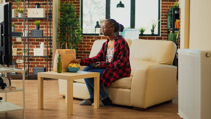 Wall Mural - African american girl receiving takeaway meal at home, preparing to watch movie on television and eat fast food in paperbag. Young cheerful woman enjoying film on tv, drinking beer.