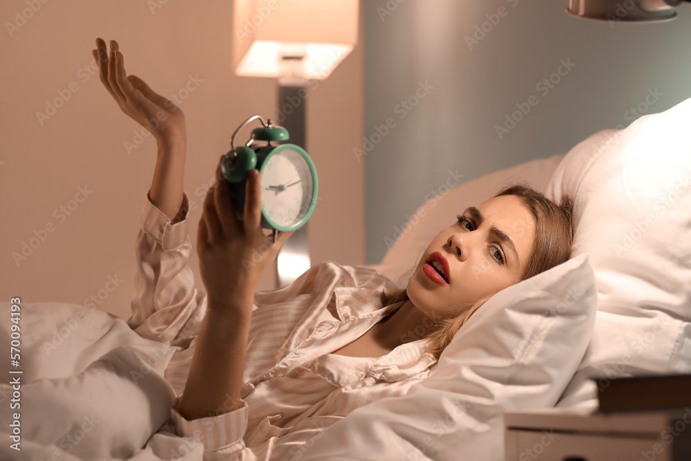 Upset young woman with alarm clock lying in bed at night