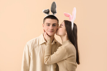 Poster - Young woman in bunny ears kissing her boyfriend on beige background. Easter celebration