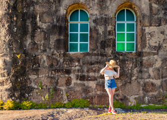 Woman tourist wearing hat and gracefully posing in front of old stone wall with two green glass windows.