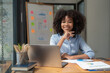 © Songsak C - Smiling African American businesswoman working on a laptop sitting at her desk in a modern office.
