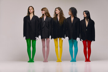 Wall Mural - Studio shot of young stylish beautiful fashion models in black jackets and multi colored tights posing over grey background. Concept of beauty, feminism, emotions, extraordinary fashion