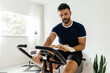 © kleberpicui - Man working out on exercise bike while reading a book at home