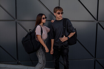 The fashion couple of girl and boy teenagers dressed black t-shirt, jeans, sunglasses, watches and bagback walking in the street at the background grey wall