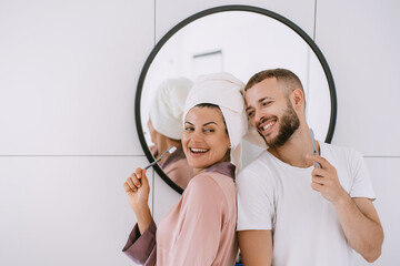 Cheerful caucasian young couple at bathroom brushing teeth toothy smiling cuddling. Newlyweds enjoying honeymoon, at hotel room. Pretty hispanic girl with towel on head having fun with boyfriend.