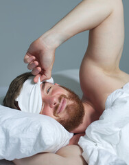 Wall Mural - Young happy handsome sleepy man in the bed in bedroom at home in the morning lying under white blanket, enjoy resting, sleep well in night mask on face, eyes. Vertical photo 