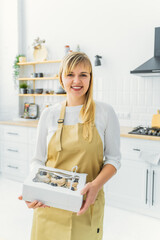 a blonde pastry chef in a bright kitchen in an apron holds a white box with muffins in her hands, de