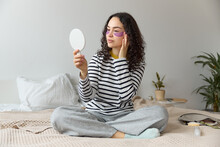 Horizontal Close Up Of Young Beautiful Dark Curly Haired Cheerful Beautiful Woman Applying Gel Purple Eye Patches Looking In A Pocket Mirror While Sitting On A Bed N A Modern Interior Room At Home. 