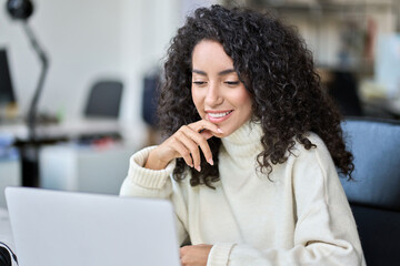 Young happy latin business woman company worker sitting in office working on laptop tech. Smiling female professional sales or marketing manager using corporate services on computer browsing internet.