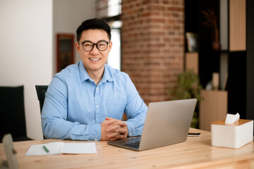 Portrait of happy middle aged asian businessman sitting at desk with laptop in home office and smiling, free space