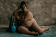 portrait of a nude plus size woman sitting on the ground leaning on a box beside a blue vase