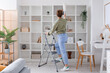 Young woman on stepladder cleaning bookshelf with duster at home
