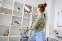 Young Woman On Stepladder Cleaning Bookshelf With Duster At Home