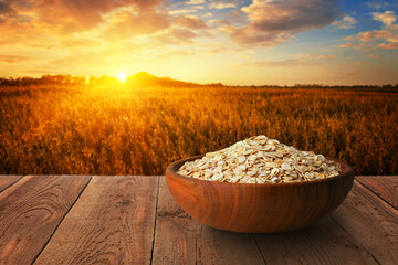 Wall Mural - oat flakes in bowl on table with ripe cereal field on sunset as background