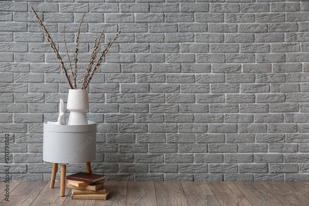Vase with willow branches on table and books near grey brick wall