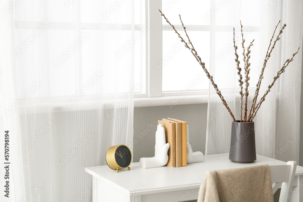 Vase with willow branches, books and alarm clock on table in light room