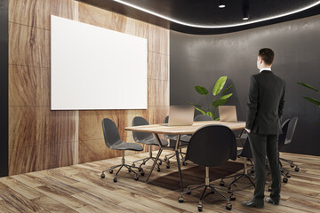 Poster - Back view of businessman standing in modern wooden and black meeting room interior with empty white mock up banner, equipment, furniture and decorative plants. Commercial workspace concept.