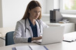 © fizkes - Positive focused pretty doctor woman typing on laptop at work table in clinic office. Physician, female practitioner in white coat, stethoscope using computer for online communication