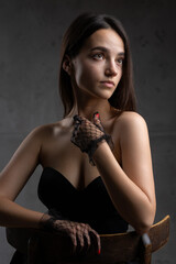 Classic dark studio portrait of a young brunette woman in black clothes who is sitting on a chair.