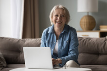 Happy Joyful Older Pensioner Woman Using Posing At Computer, Sitting On Sofa At Home, Using Laptop For Distant Studying, Online Communication, Looking At Camera, Smiling, Laughing. Female Portrait
