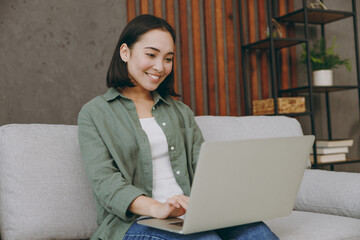 Young fun IT woman of Asian ethnicity wears casual clothes hold use work on laptop pc computer sits on grey sofa couch stay at home hotel flat rest relax spend free spare time in living room indoor.