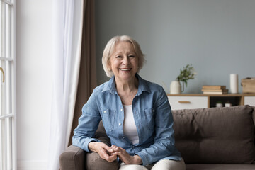 joyful positive senior lady sitting on sofa, smiling, laughing, looking at camera, enjoying retireme