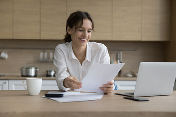 Happy freelance professional working with document from home, sitting at kitchen table, laptop, calculator, reading legal paper, smiling, laughing, getting good news