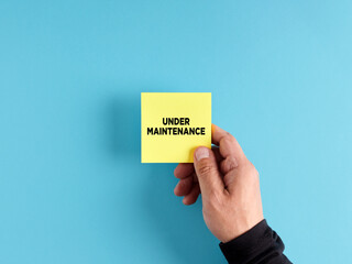 Wall Mural - male hand holds a yellow sticky note paper with the message under maintenance.