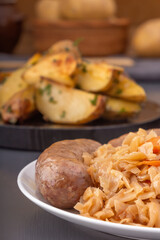 Braised cabbage - bigos and grilled sausage in a white porcelain plate against the background of rustic potatoes and pickled vegetables. Close-up