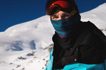 The skier looks at the camera before starting to ski. Man enjoys vacation in winter season. Portrait of a man in a balaclava and ski goggles against the backdrop of a snowy mountain