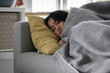 © DimaBerlin - Sick Asian woman sleeping resting on sofa under blanket at home, recovering from flu. Sleepy weak female lying on couch suffering from fever and chills during sickness, having symptoms of Influenza