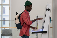 Young African Student Guy With Backpack Standing In Library Using Self-service Terminal To Borrow Or Return Books, Searching Literature Browsing Catalogue, Touching Screen Of Self-checkout Kiosk