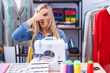 Poster - Blonde woman dressmaker designer using sew machine peeking in shock covering face and eyes with hand, looking through fingers with embarrassed expression.