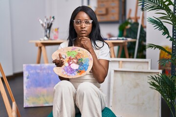 Wall Mural - African young woman holding painter palette serious face thinking about question with hand on chin, thoughtful about confusing idea