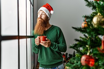 Canvas Print - Young redhead man drinking coffee standing by christmas tree at home