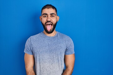 Wall Mural - Middle east man with beard standing over blue background sticking tongue out happy with funny expression. emotion concept.