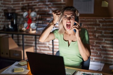 Wall Mural - Young beautiful woman working at the office at night speaking on the phone angry and mad raising fist frustrated and furious while shouting with anger. rage and aggressive concept.