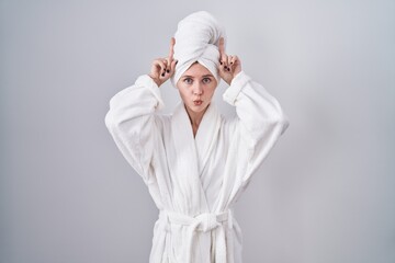 Poster - Blonde caucasian woman wearing bathrobe doing funny gesture with finger over head as bull horns