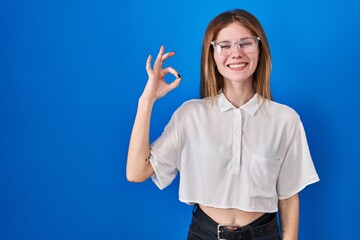 Poster - Beautiful woman standing over blue background smiling positive doing ok sign with hand and fingers. successful expression.