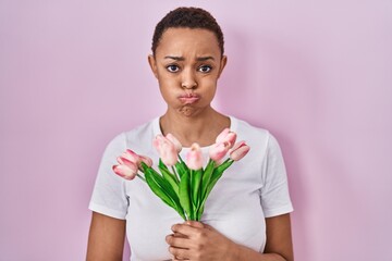 Sticker - Beautiful african american woman holding bouquet of pink tulips flowers puffing cheeks with funny face. mouth inflated with air, catching air.