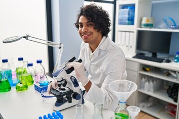 Sticker - Young hispanic man wearing scientist uniform using microscope at laboratory