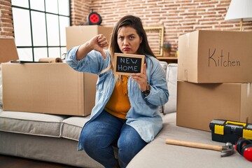 Wall Mural - Young hispanic woman holding blackboard with new home text with angry face, negative sign showing dislike with thumbs down, rejection concept