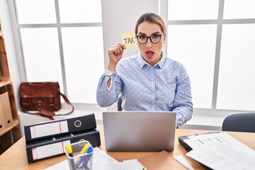Wall Mural - Young hispanic business woman at the office calculating taxes scared and amazed with open mouth for surprise, disbelief face